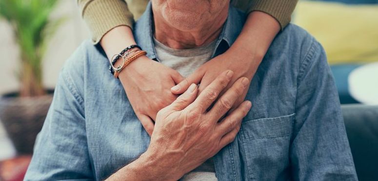 Close-up of an elderly person being embraced from behind, with both individuals' hands resting together on the elder's chest.