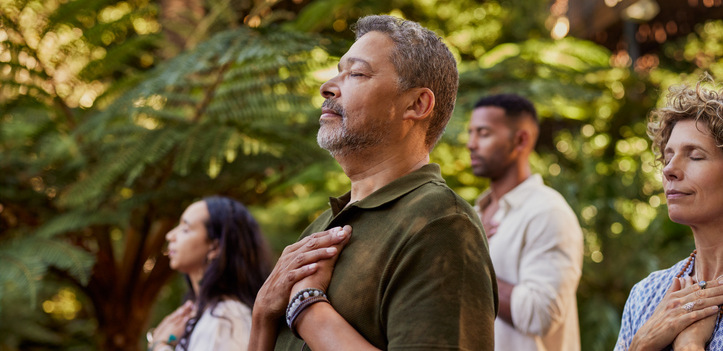 Group of multiethnic people standing outdoors with eyes closed and hands on their hearts during a meditation session. Multiracial adults practicing gratitude and mindfulness together. Mature latin man connecting to inner peace and presence at wellness retreat center in nature.