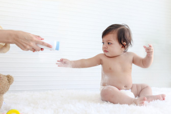 Portrait of happy smiling toddle baby girl sit on fluffy white rug with mother hand hold dusting powder bottle, mom apply talcum powder on body of little cute kid daughter child, childhood care family