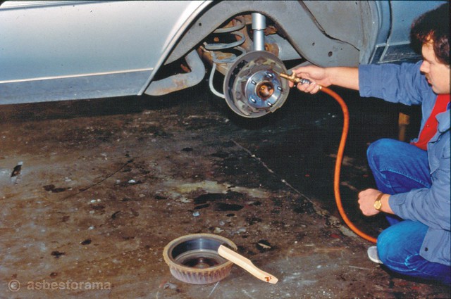 Mechanic uses air hose to remove asbestos on brakes.