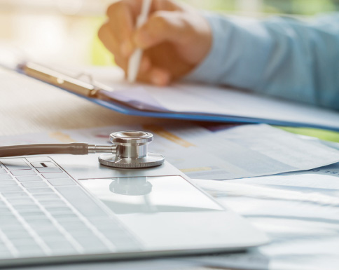 A doctor taking notes on clipboard in front of a stethoscope and laptop